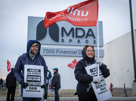Two Unifor members outside on a picket line wearing On Strike placards. One is carrying a Unifor flag.