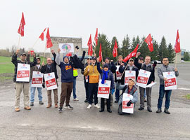 A group of people holding red and white Unifor 'On Strike' signs and some waving red Unifor flags.